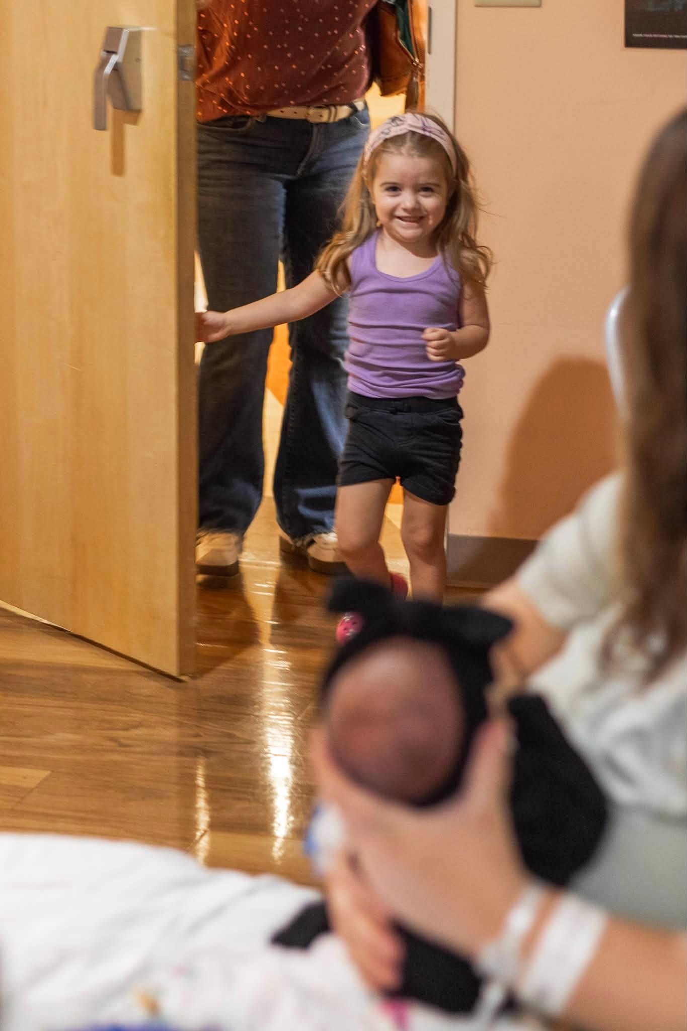 A smiling child enters a room as a newborn is held nearby
